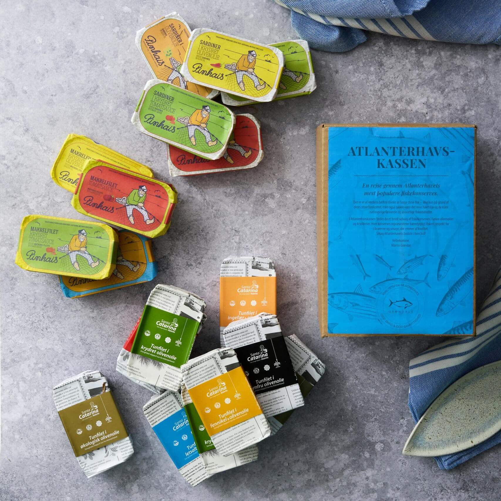 Grøndals Atlanterhavskassen med bæredygtigt fisket fisk og skaldyr som portugisisk makrel er udstillet i farverige dåser på en grå overflade ved siden af en blå æske med blå- og hvidstribede håndklæder i baggrunden.