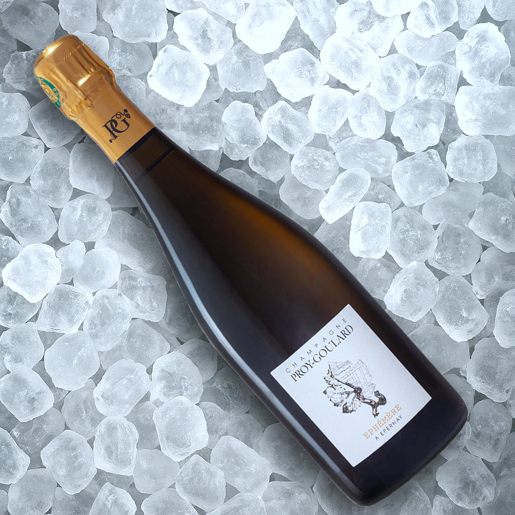 En flaske Champagne Proy-Goulard Éphémère Millésime 2019 BDN med guldfolietop og hvid etiket hviler diagonalt på klare isterninger, og på etiketten står der "Champagne Proy-Goulard.