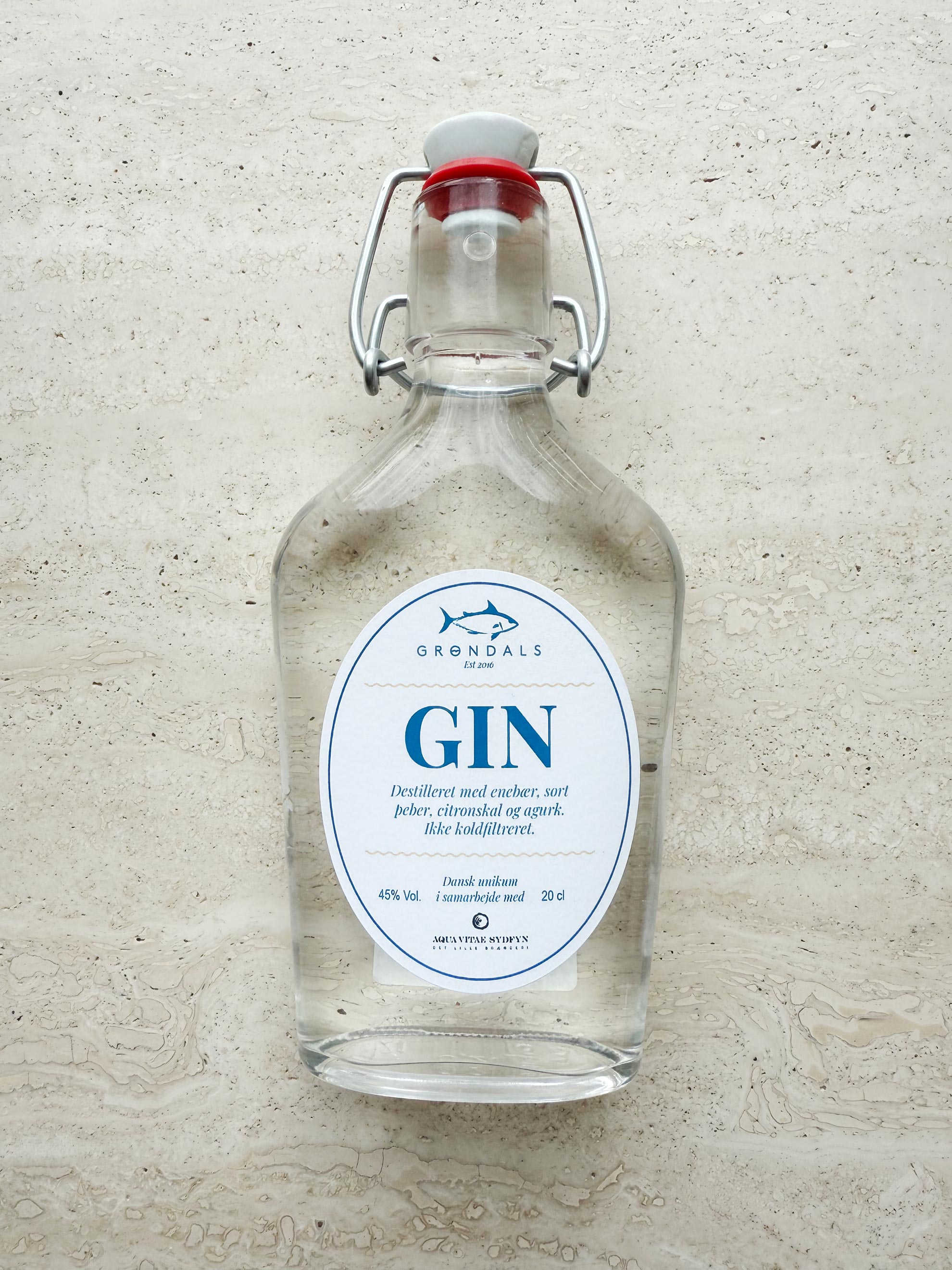 En klar Grøndals Gin - Frisk, krydret og skabt til fisk-flaske med svingprop og hvidblå etiket hviler på en tekstureret sten - en perfekt håndlavet gin til fisk fra Grøndals.