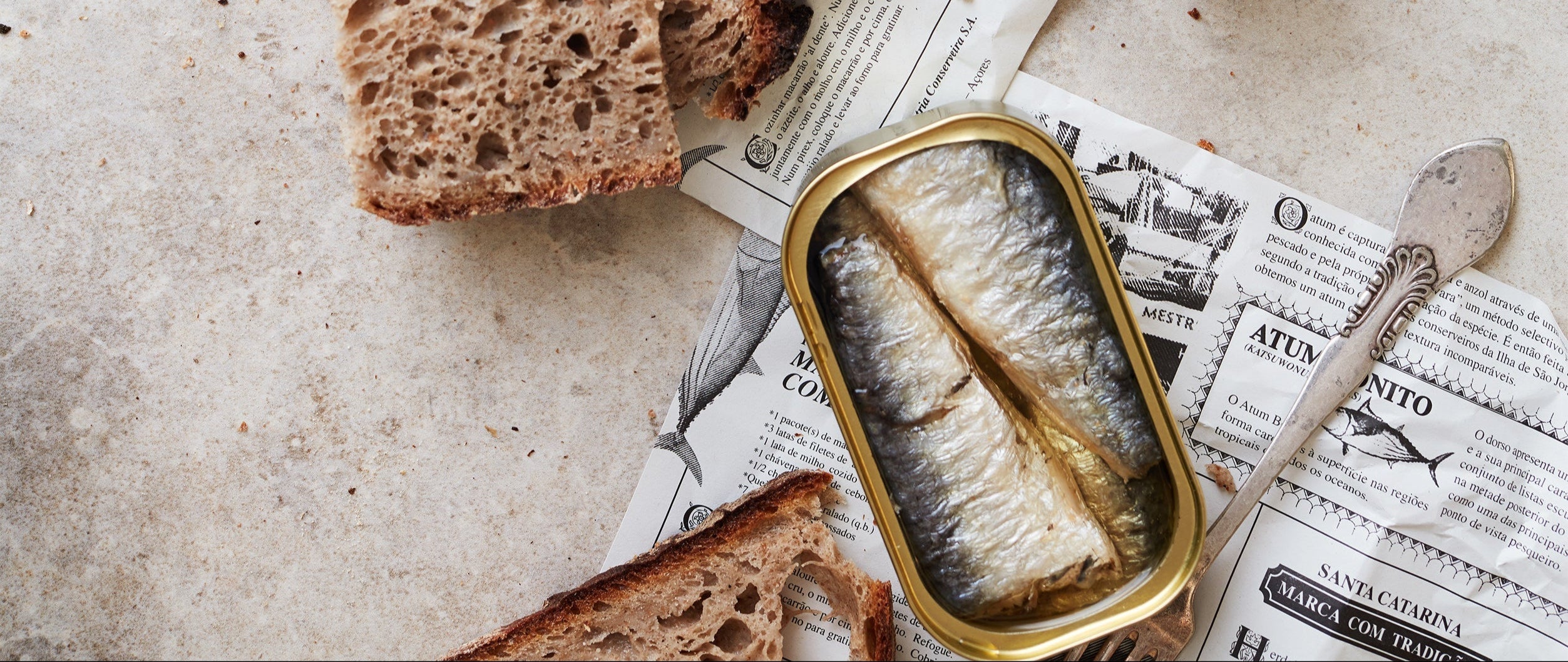 En åben dåse sardiner ligger på trykt papir med en sølvske ved siden af, omgivet af skiver af rustikt brunt brød på en lys stenoverflade.