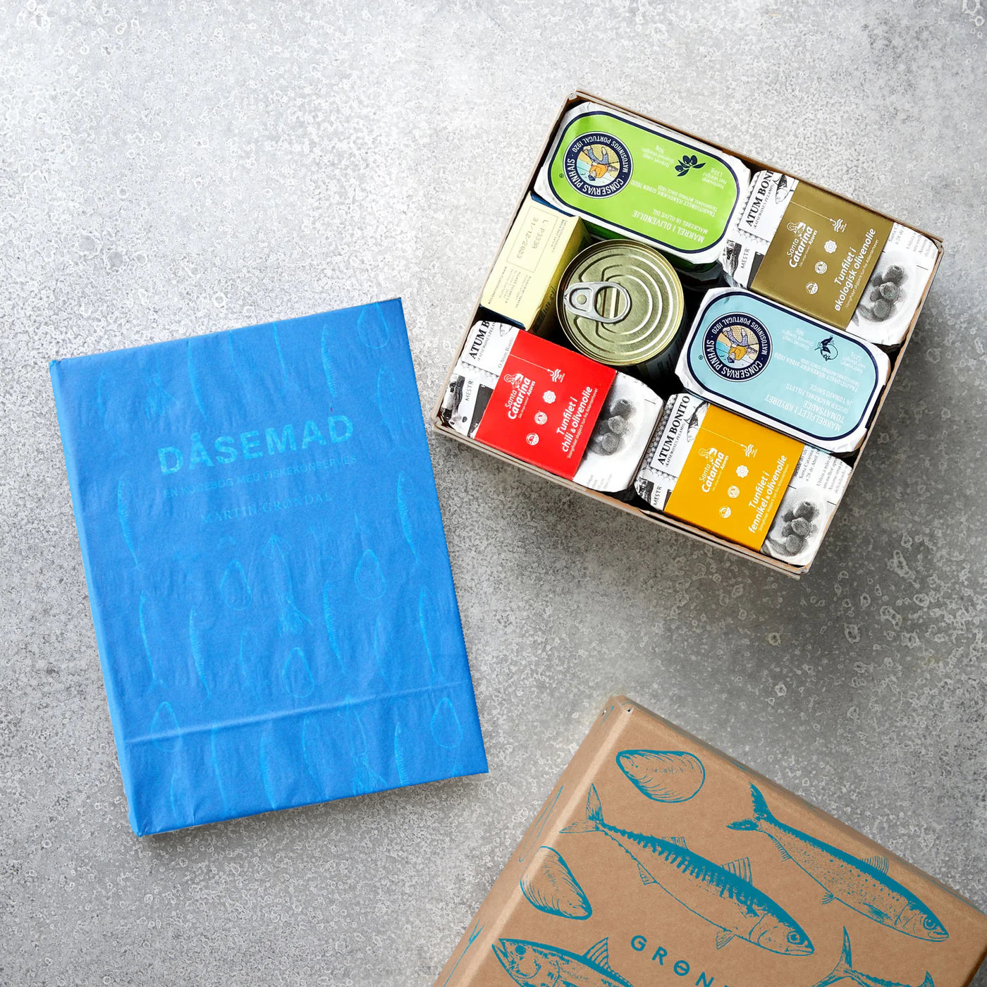 Grøndals' Smagekassen og DÅSEMAD - med en blå kogebog, en æske med forskellige fiskekonserves og en brun æske med blå fiskeillustrationer - vises på en grå flade.
