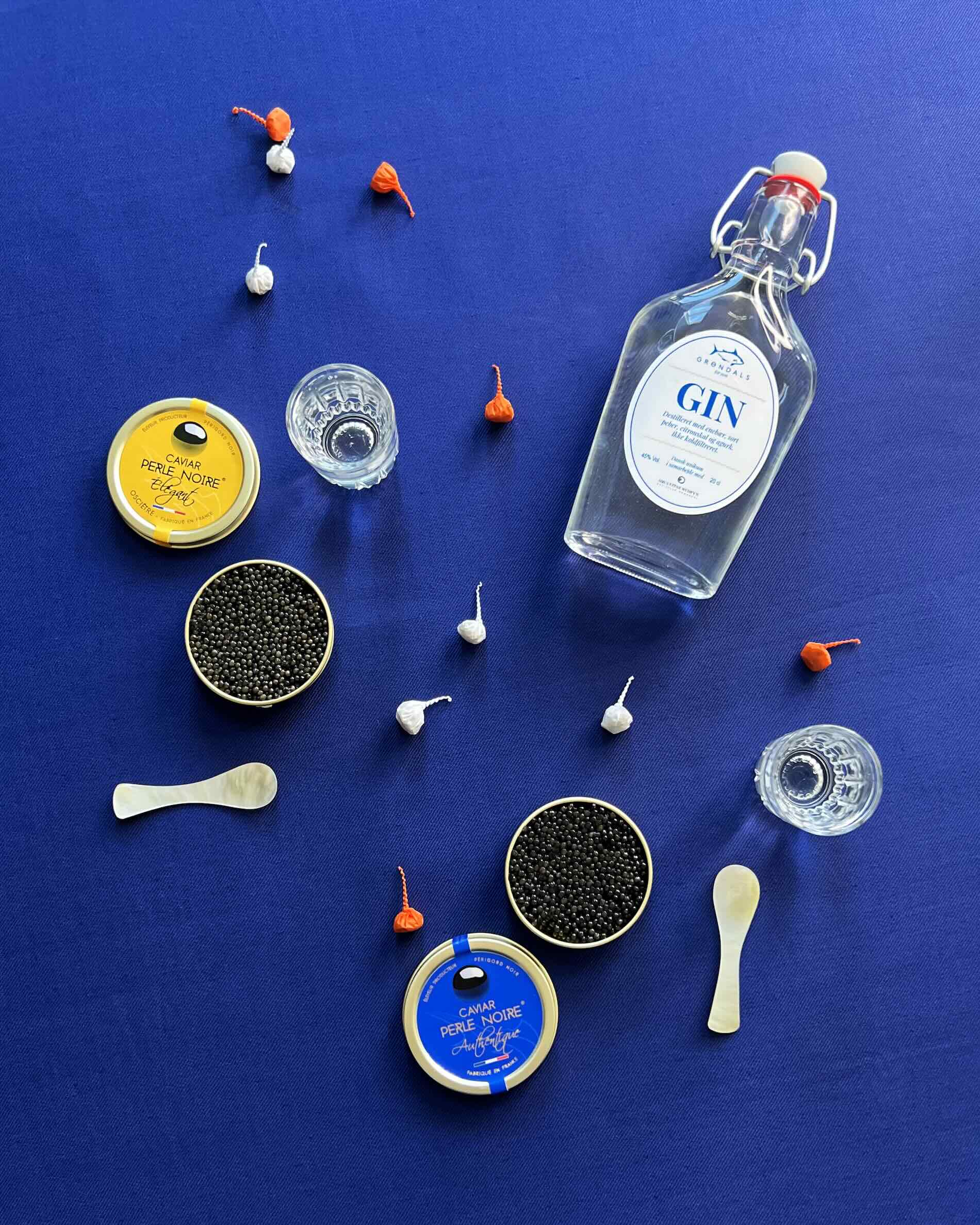En flaske gin, to dåser kaviar, tre shotglas, to små elfenbensskeer og spredte orange og hvide indpakkede bolsjer er arrangeret på en dybblå overflade.