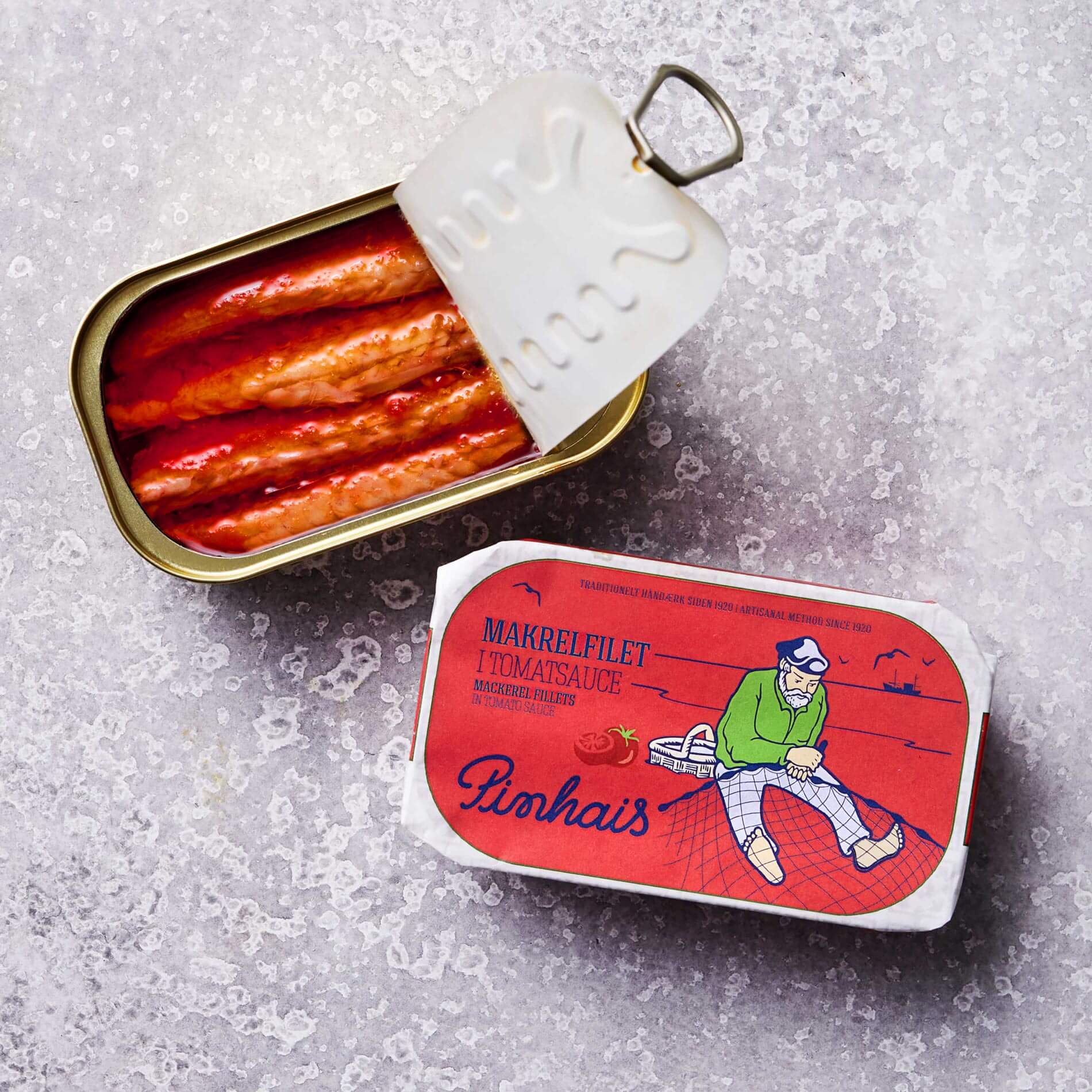 An opened tin of Makrelfilet i tomatsauce sits beside its closed packaging, displaying an illustrated fisherman in green on a red background. The tins, rich in omega-3-fedtsyrer, are placed on a textured gray surface.