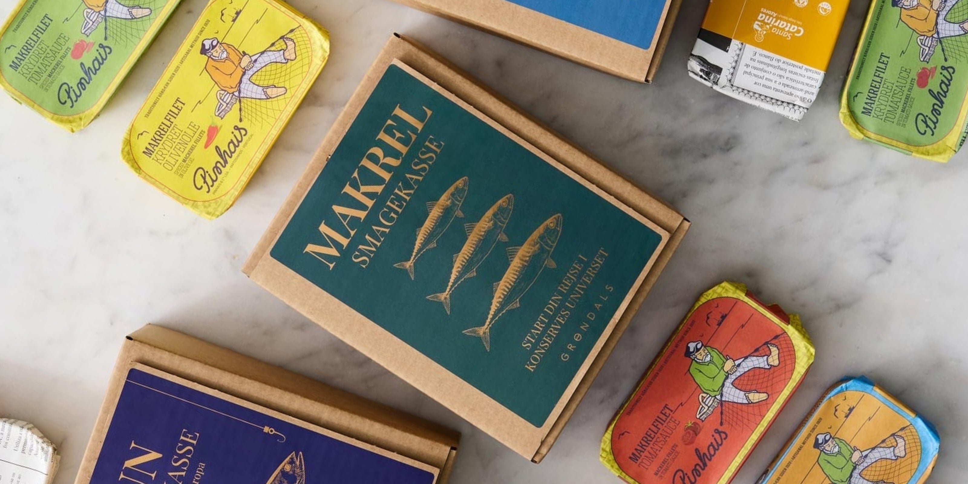 Et udvalg af farverige fiskedåser og en papkasse med påskriften Makrel Smagekasse arrangeret på en marmorflade. Dåserne har legende illustrationer af mennesker og fisk.