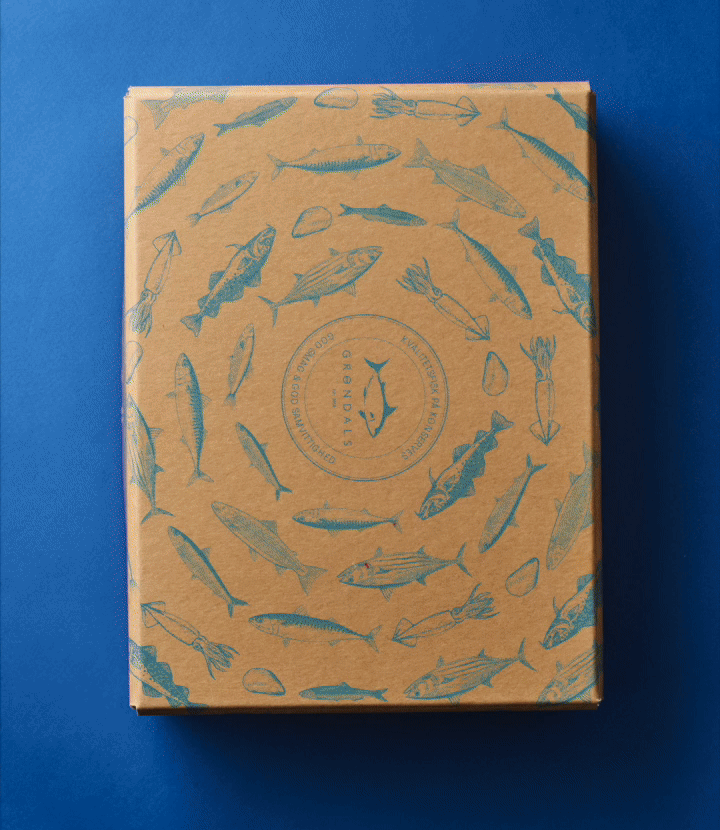 En brun æske med blå illustrationer af forskellige fisk og skaldyr på låget, arrangeret i et cirkulært mønster på en blå baggrund.