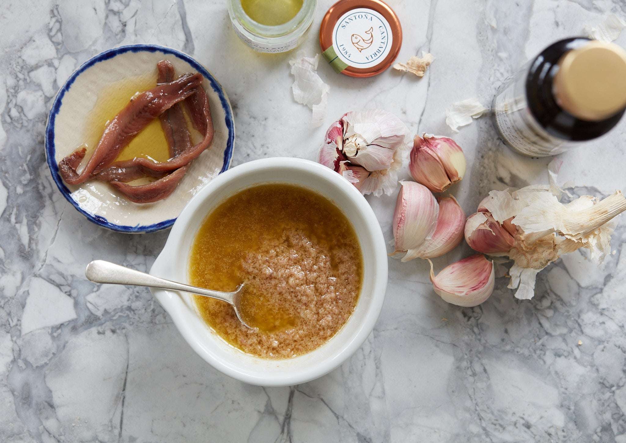 En skål med brun sovs med en ske, omgivet af Pujado Solano OUTLET | 3 x Saltede ansjosfileter i solsikke, hele hvidløgsløg, en flaske olie og en åben krukke på en marmoroverflade.