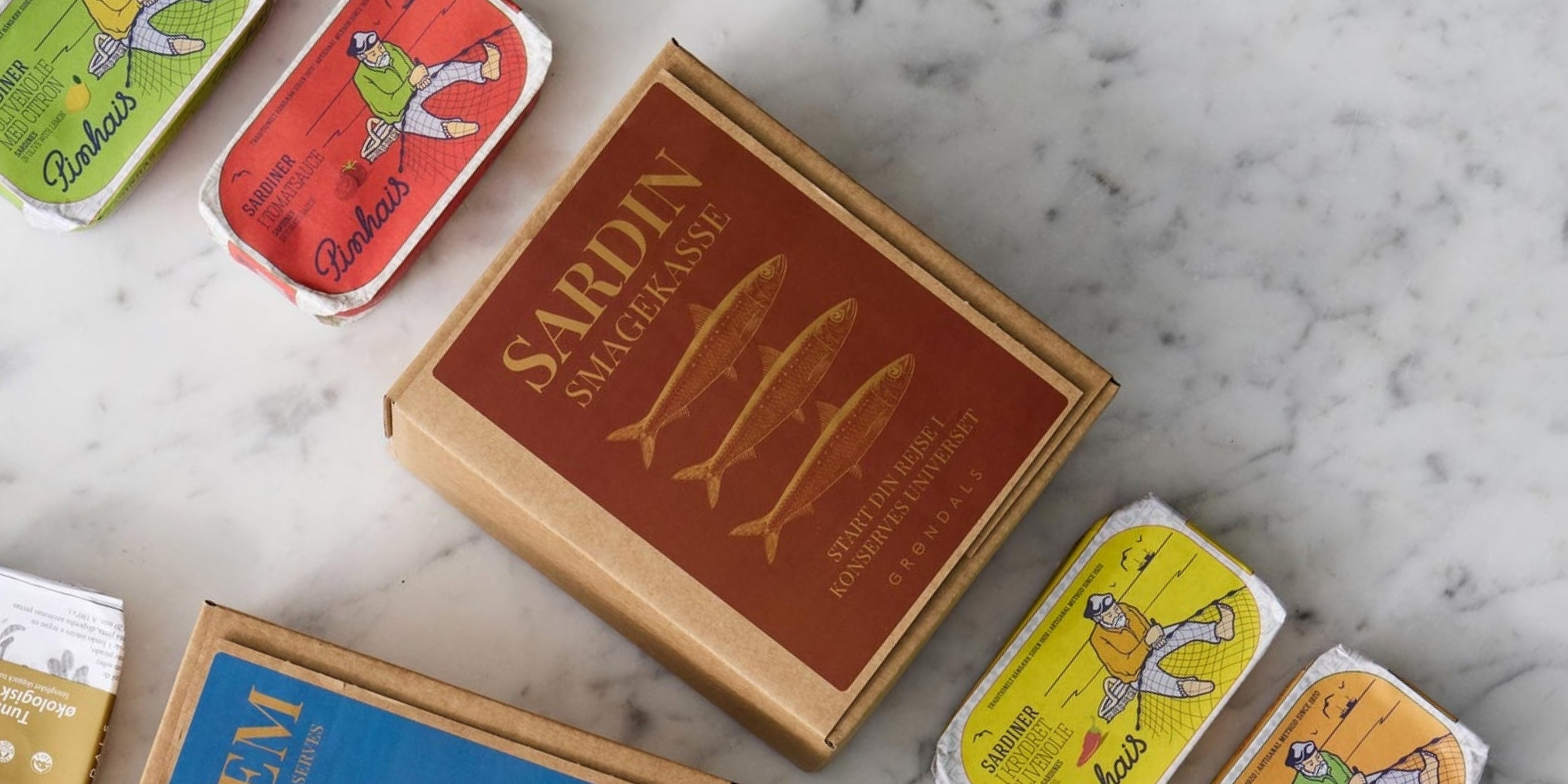 En brun æske med påskriften SARDIN SMAEGKASSE og tre fiskeillustrationer står på en marmorplade omgivet af farverige sardindåser med forskellige emballagedesigns.