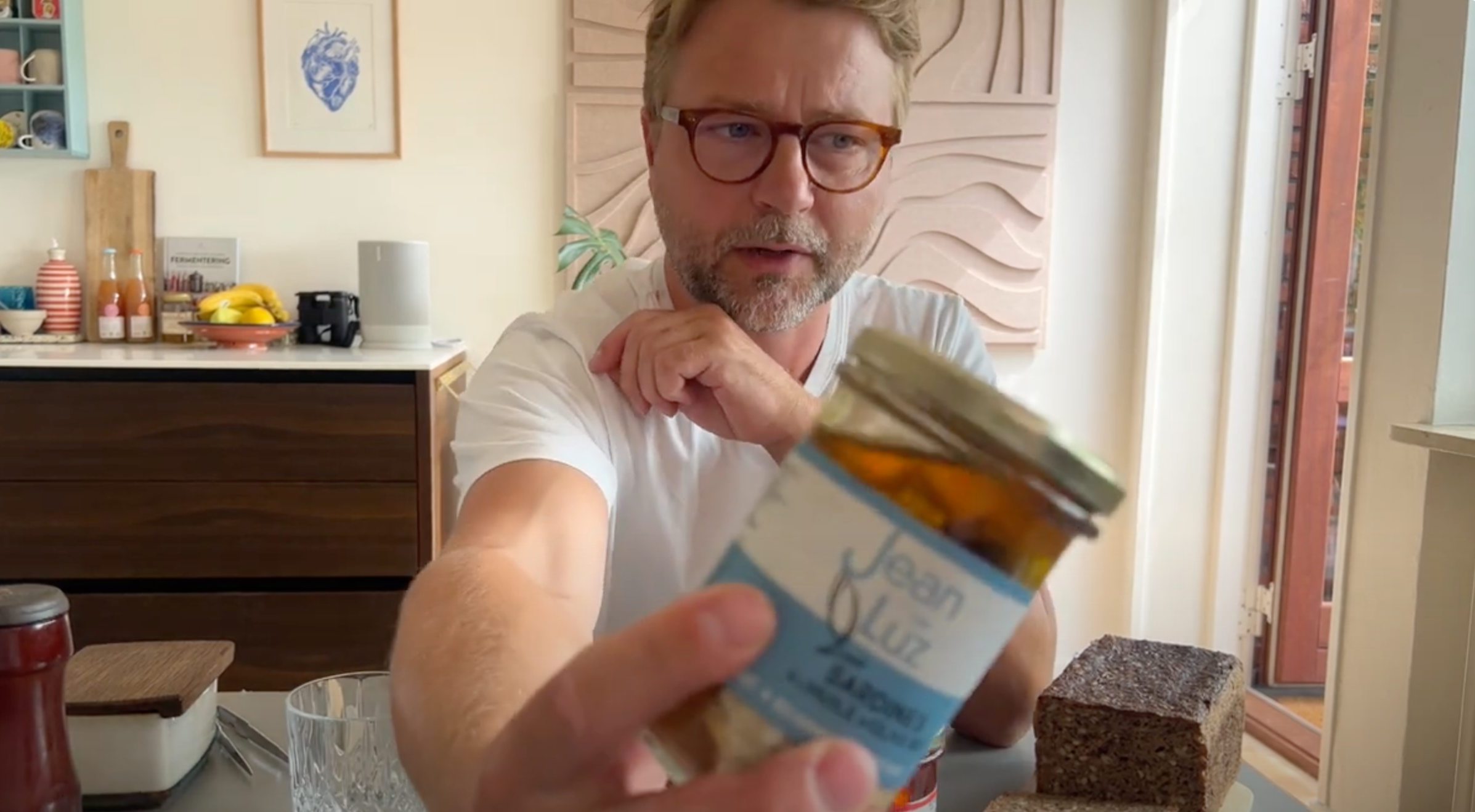 En mand med briller og hvid skjorte sidder ved et køkkenbord og holder en krukke mærket Jean Luz op mod kameraet. Brød, glas og frugt er synlige på bordet og disken bag ham.