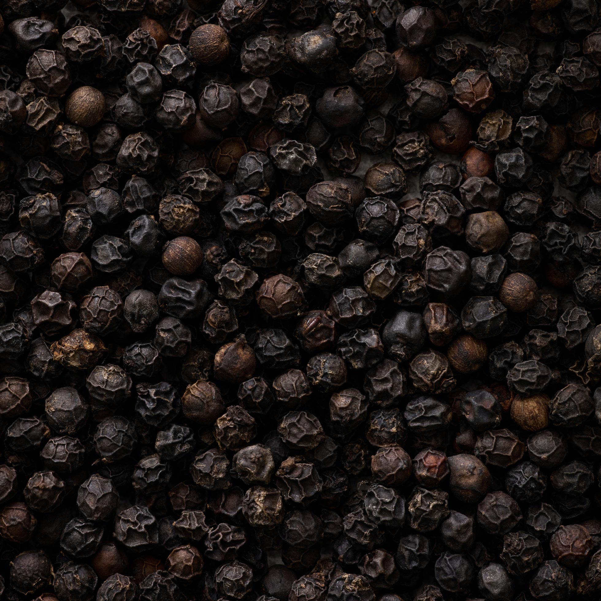 Et nærbillede af Sort Peber - i verdensklasse fra Kandy Spices viser srilankanske peberkorn med ru, teksturerede overflader og mørkebrun til sort farve, der fylder hele billedet.