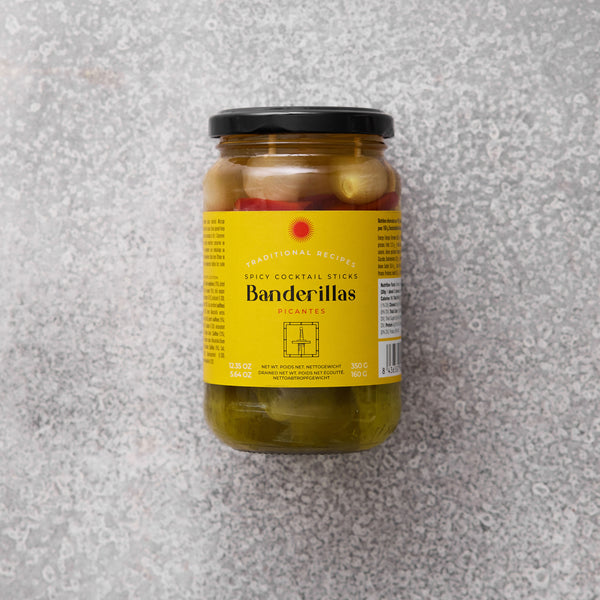 Et glas med Grøndals Spicy Banderillas cocktailpinde med sort låg og gul etiket med rød tekst står på en lysegrå struktureret overflade - disse syltede grøntsager er den perfekte snack.