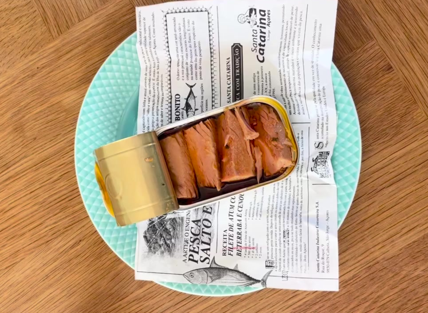 En åben dåse tun i olie ligger på trykt papir oven på en lyseblå tekstureret tallerken, der er placeret på en træplade. Tunen er skåret i stykker, og dåselåget er delvist foldet tilbage.