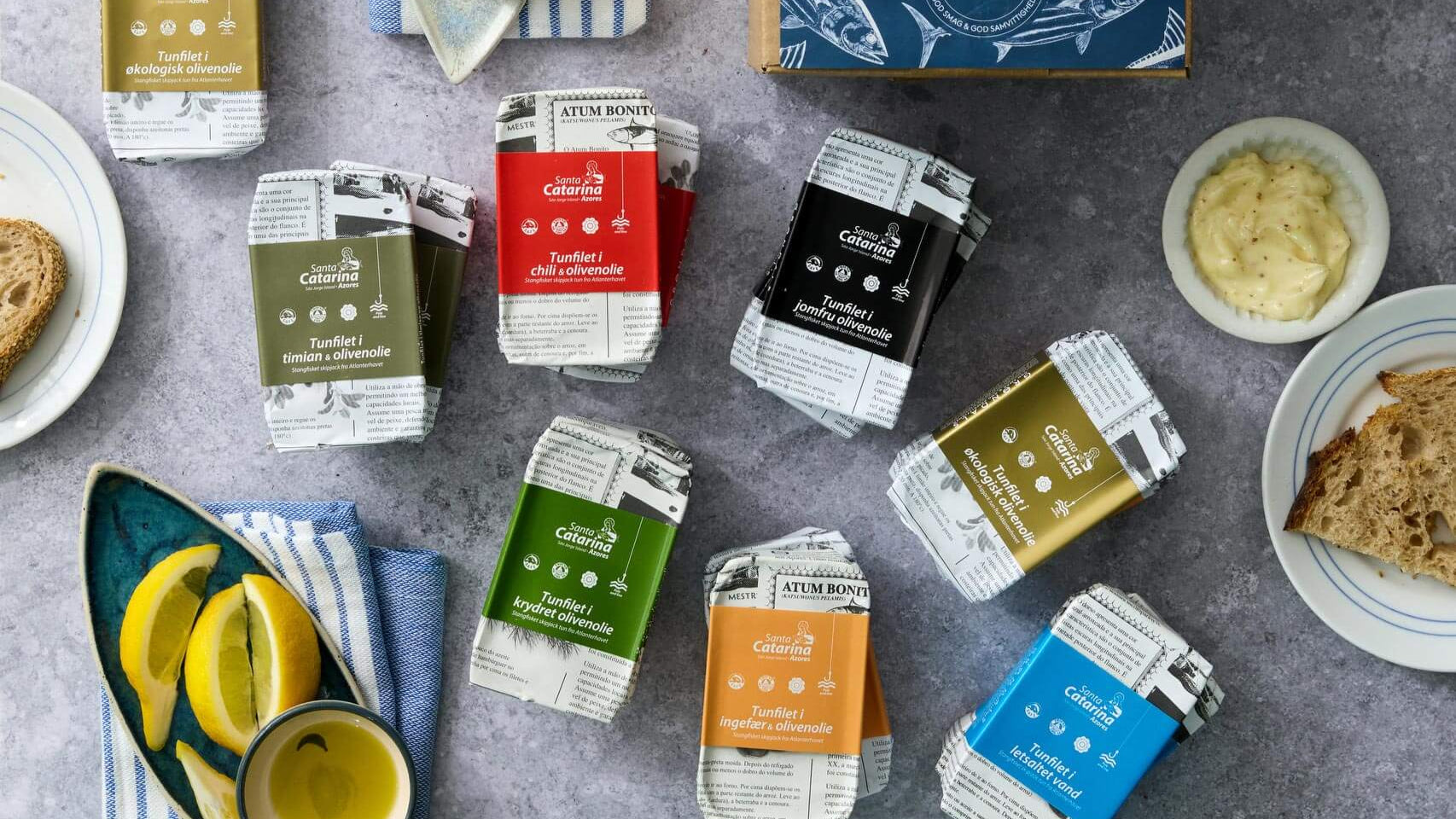 Assorterede Grøndals Atlan Smolt tunfilet i økologisk olivenolie dåser i BPA-fri dåser er arrangeret omkring en blå Tunfiletkasse på en grå overflade, med brød, mayo, citronskiver og et stribet håndklæde i nærheden.