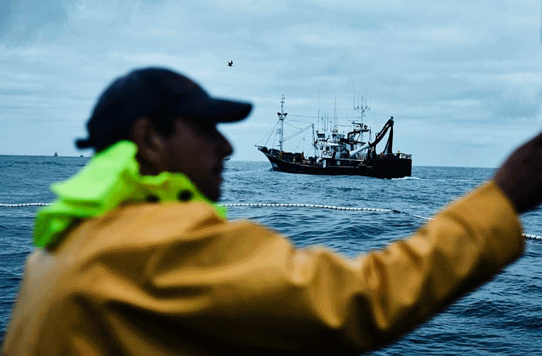 En person i en gul regnfrakke står på en båd på havet med et fiskefartøj i baggrunden under en overskyet himmel.