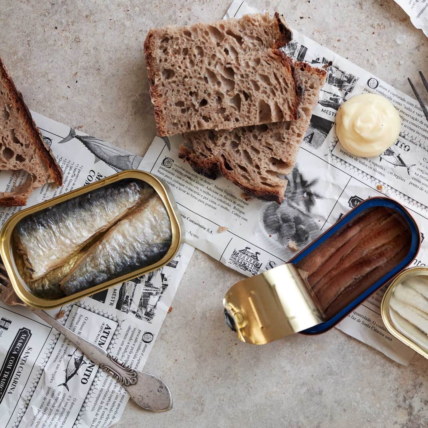 To åbnede dåser med fisk, skiver af rustikt brød og en klat mayonnaise er arrangeret på avisark på en lys overflade. En smørkniv ligger i nærheden.