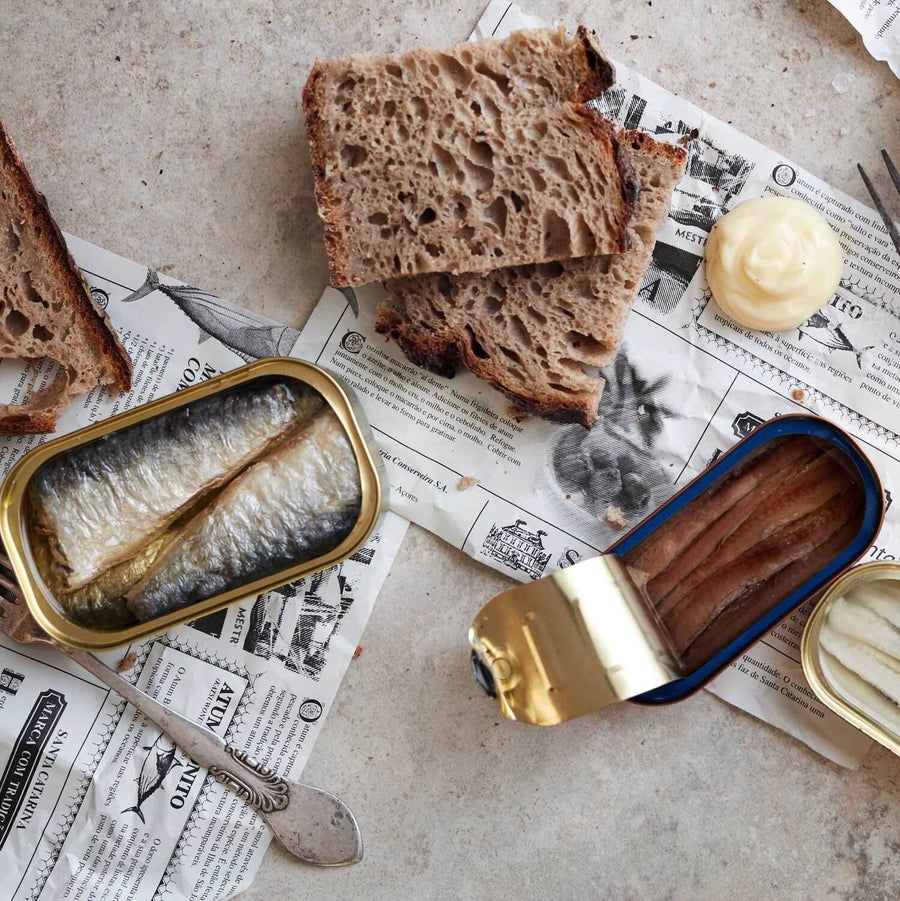 To åbnede dåser med fisk, skiver af rustikt brød og en klat mayonnaise er arrangeret på avisark på en lys overflade. En smørkniv ligger i nærheden.
