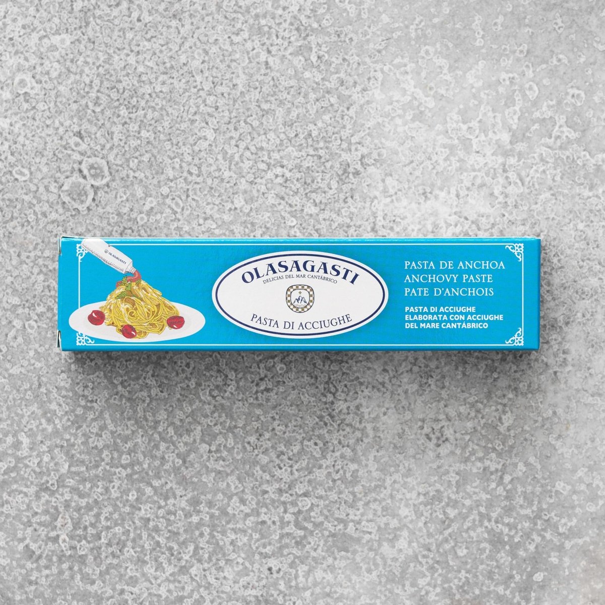 En blå rektangulær æske med Olasagasti Ansjospuré med olivenolie (60 g) ligger på en struktureret grå overflade og viser pasta med velsmagende ansjoscreme, tomater og flersproget information om bæredygtige ansjoser.
