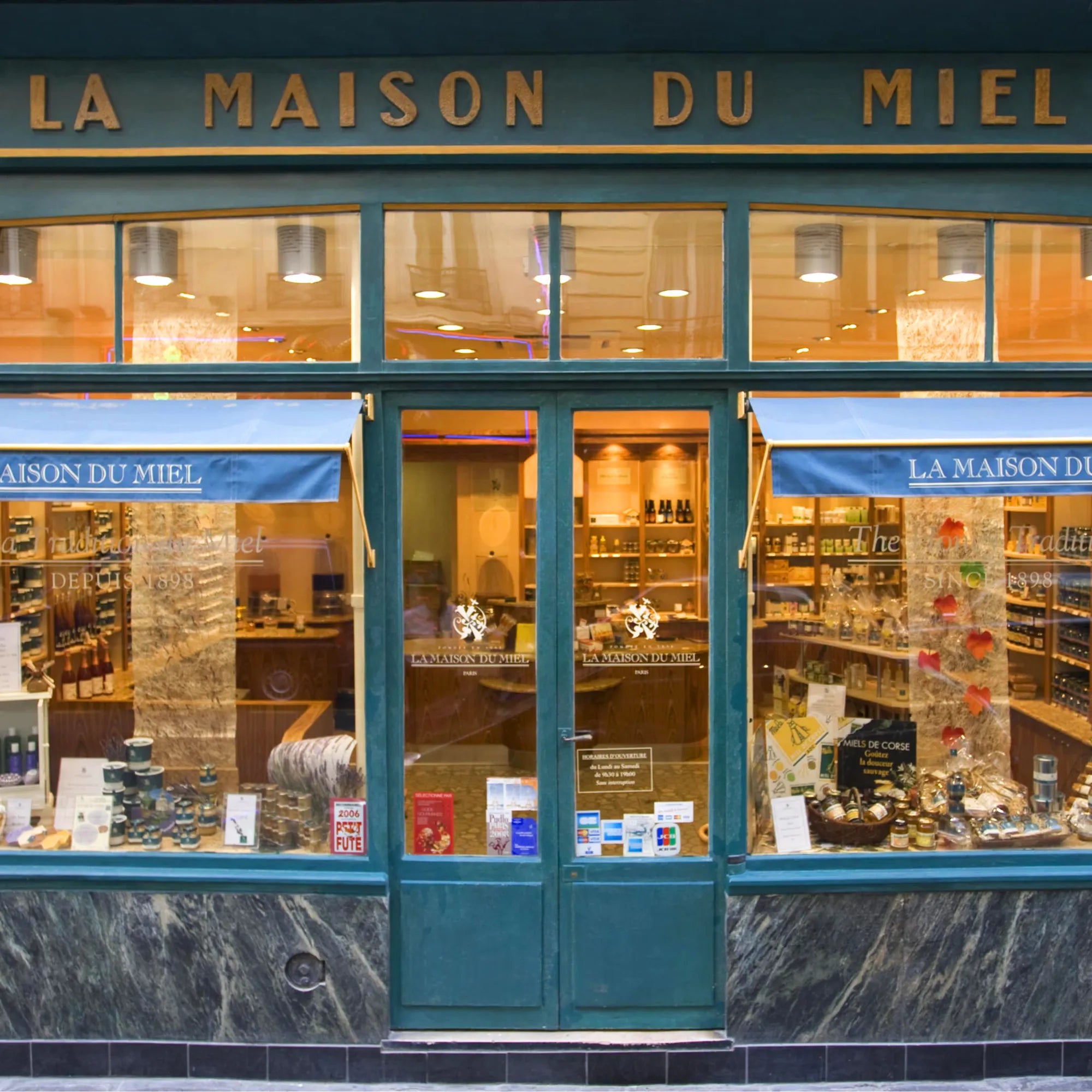 Facade af butikken “La Maison du Miel” med store vinduer, der afslører hylder fyldt med honning og andre produkter. Butikkens navn er skrevet med store bogstaver øverst på facaden, og blå markiser pryder vinduerne. Billedet er forbundet med Grøndals.