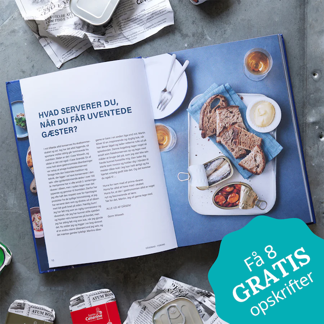 En åben kogebog med dansk tekst ligger på en blå flade, omgivet af dåser med fisk, rugbrødsskiver, smør og citron. Krøllede aviser og et blågrønt skilt med teksten Få 8 GRATIS opskrifter er også synlige.