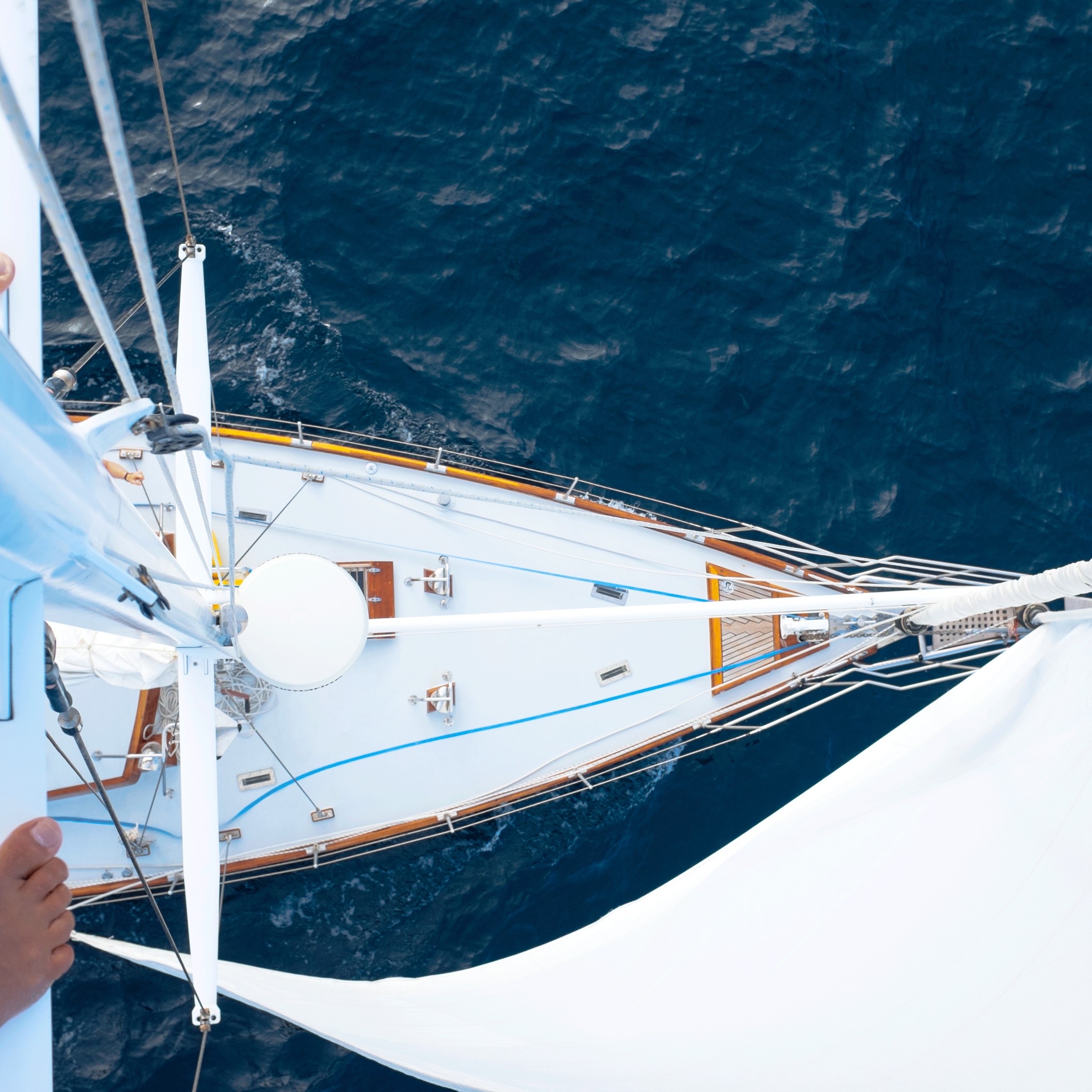 Et vue ovenfra af en sejlbåd på dybt blåt vand, der viser stævnen, det hvide dæk, riggen og sejlet. En persons bare fod er synlig på venstre side af bådens mast.