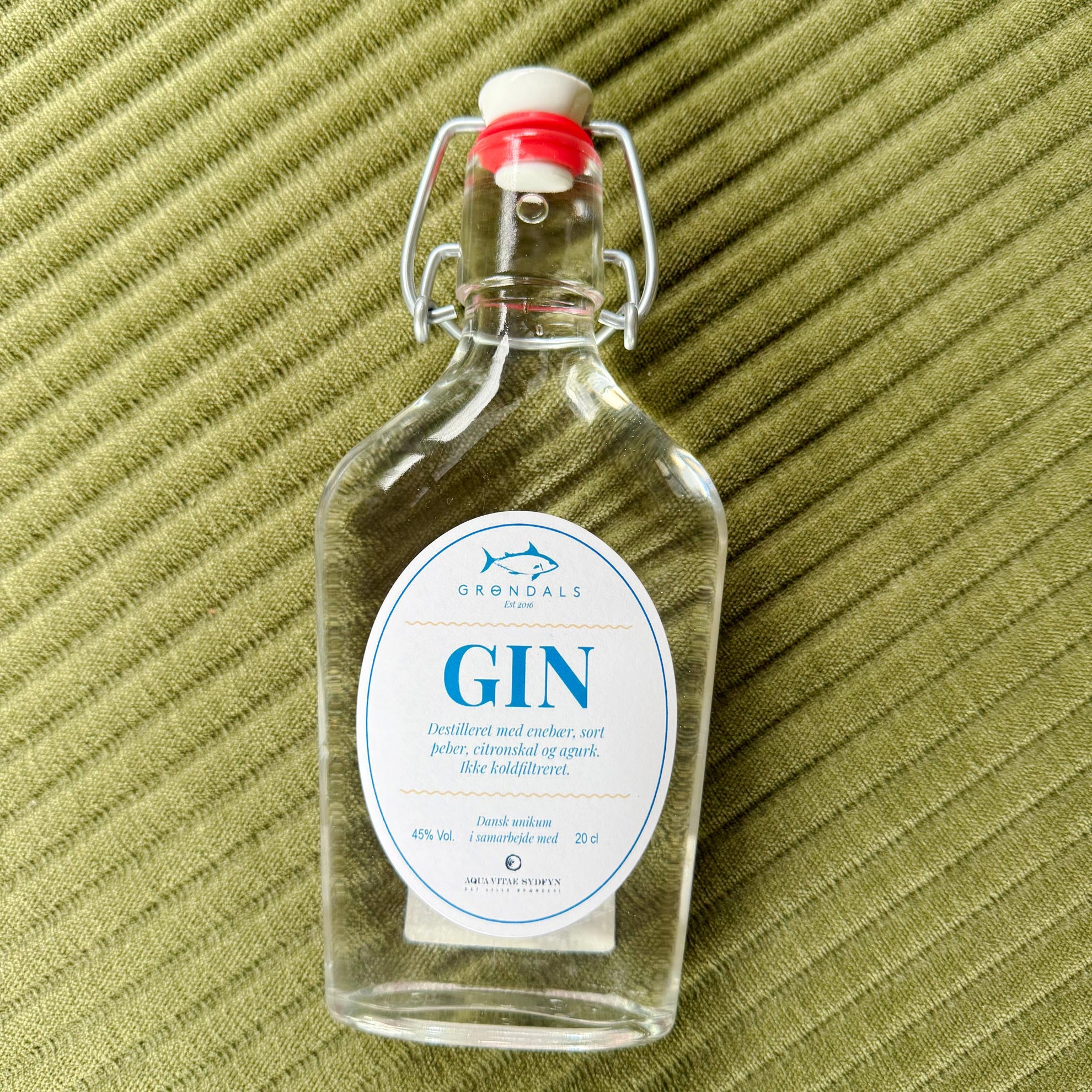 En klar glasflaske med Grøndals Gin - Frisk, krydret og skabt til fisk står på struktureret grønt stof. Den har svingprop, hvid etiket med blå tekst og fiskelogo - Grøndals' håndværksmæssige gin, der er perfekt som gin til fisk.