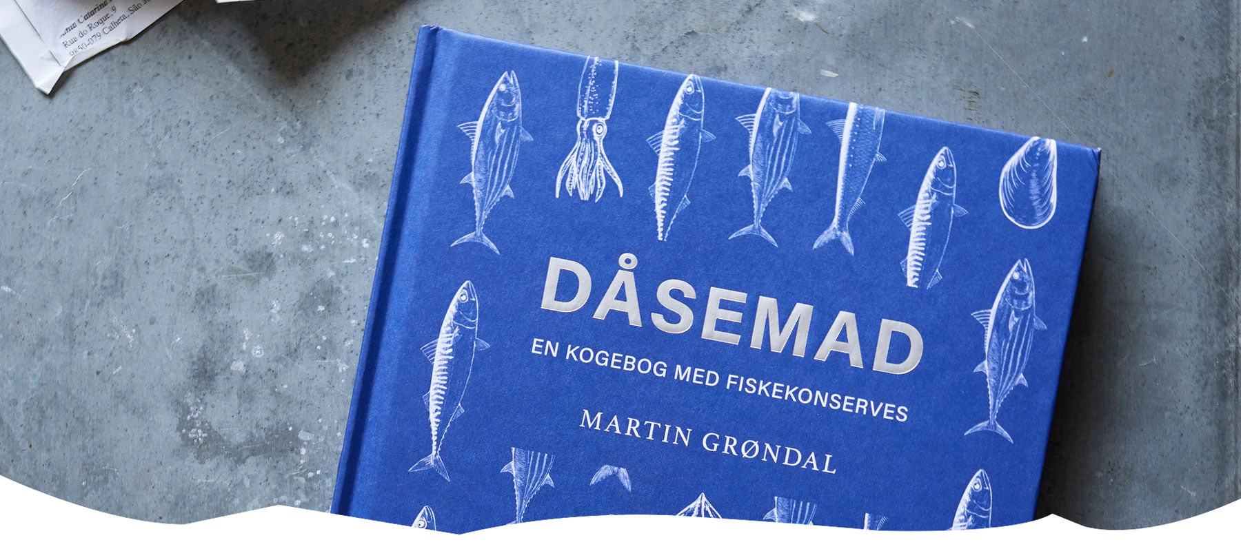 En blå kogebog med titlen Dåsemad: En kogebog med fiskekonserves af Martin Grøndal har hvide illustrationer af forskellige fisk og skaldyr på omslaget. Bogen ligger på en grå overflade.
