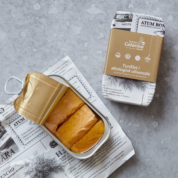 En åbnet dåse med tunfileter i olie ligger på avislignende papir ved siden af emballagen, der er mærket Santo Catarina med guld og hvidt design og tekst på et andet sprog. Baggrunden er en grå stenoverflade.