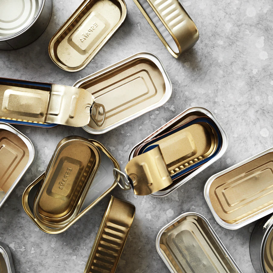Adskillige åbnede og uåbnede rektangulære blikdåser med pull-tab-låg ligger spredt på en lysegrå, marmoreret overflade. Dåserne er for det meste guld- eller sølvfarvede.