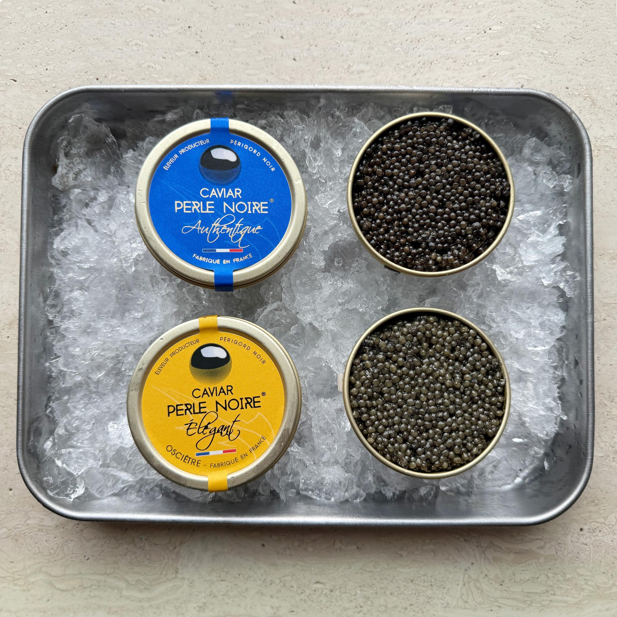 En metalbakke med is indeholder Grøndals' Kaviar-duo - L'Élégant (Oscietre) 30 g og L'Authentique (Baerii) 30 g - med to åbne dåser, der afslører små, mørke perler, og to lukkede dåser med blå og gule etiketter.