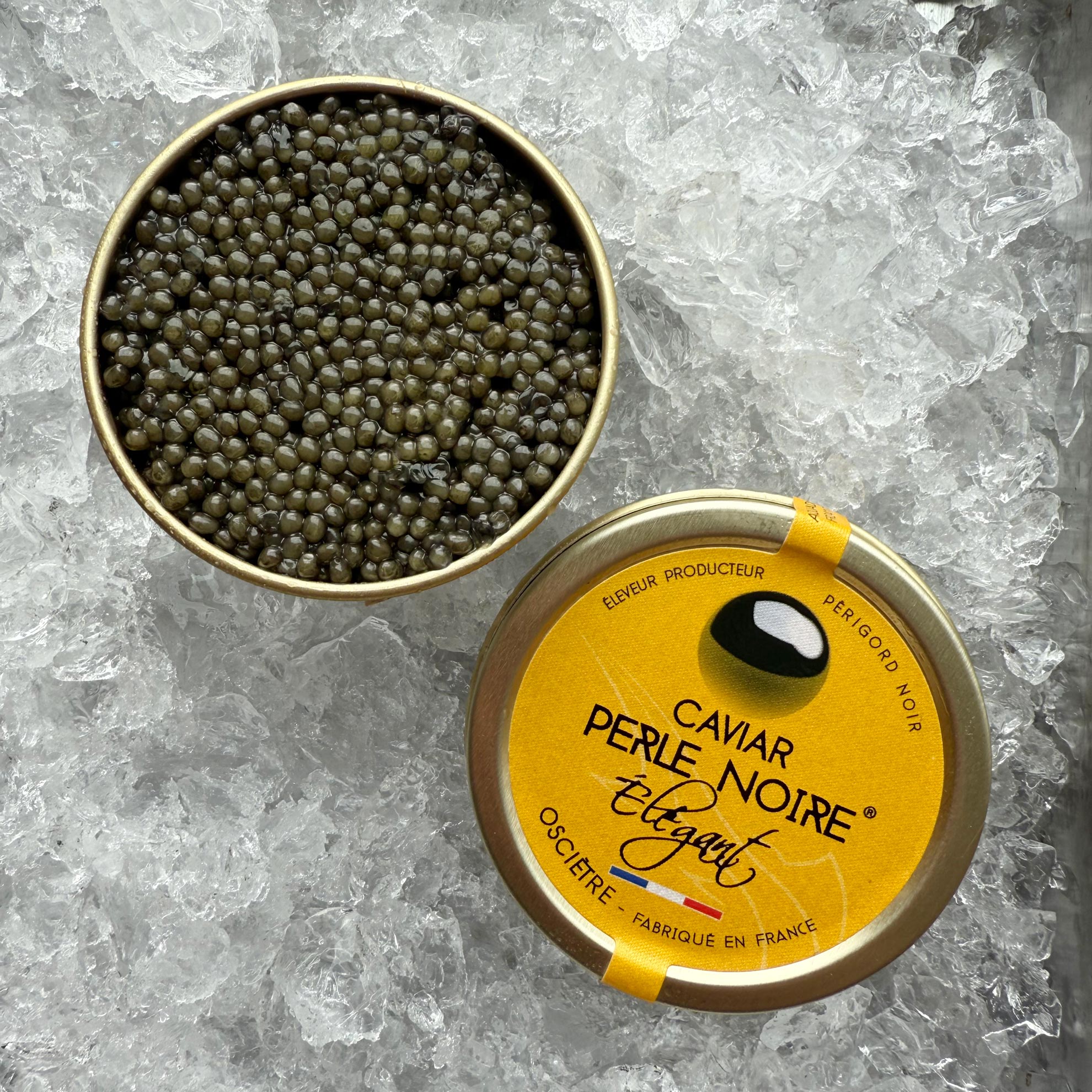 En dåse Grøndals L'Élégant (Oscietre) 30g - Den raffinerede luksus hviler på is og afslører blanke, sorte perler. Låget, der er mærket på fransk og engelsk, ligger ved siden af den åbne dåse.