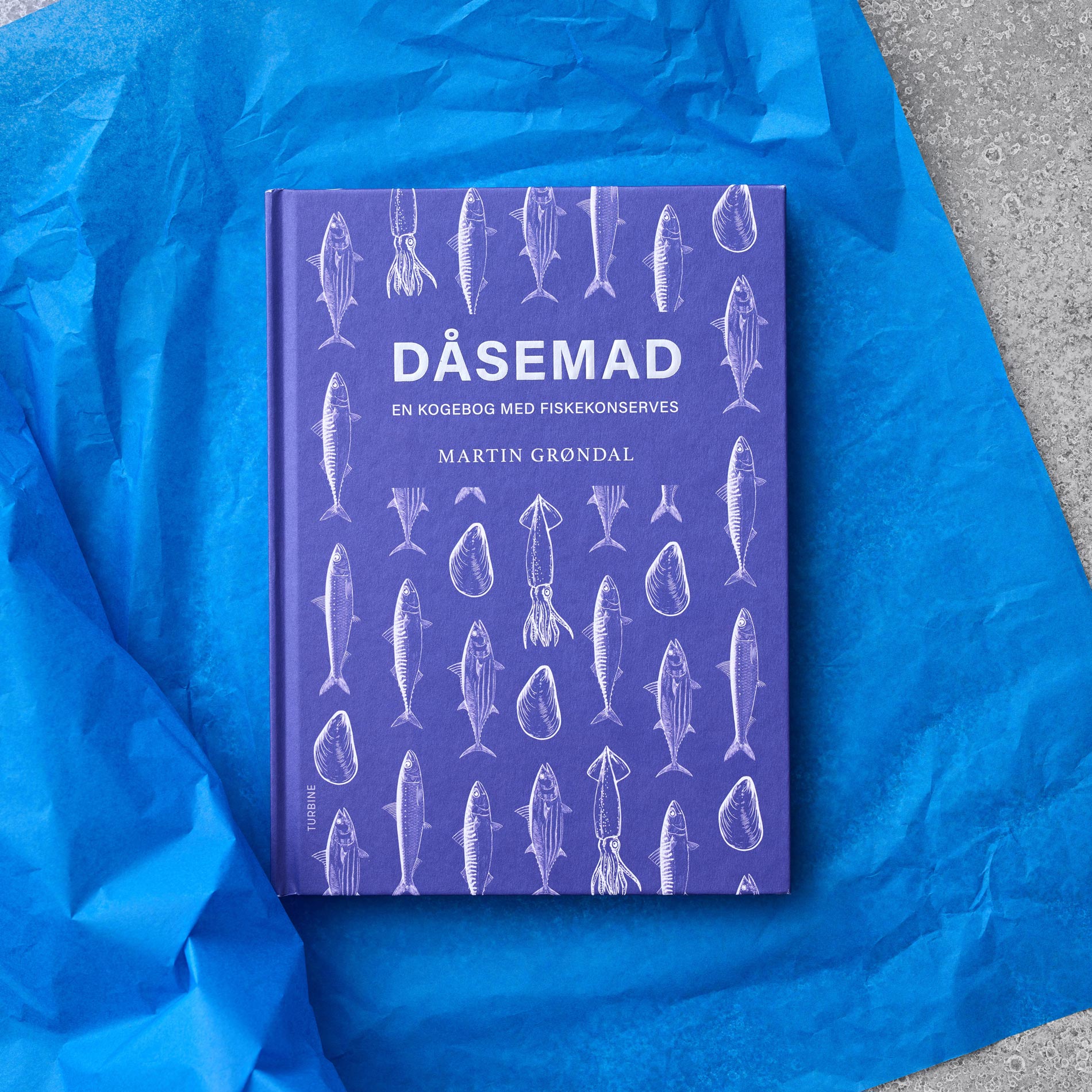 En lilla kogebog med titlen Dåsemad: En kogebog med fiskekonserves af Martin Grøndal er placeret på blåt silkepapir med hvide illustrationer af fisk og skaldyr på omslaget.