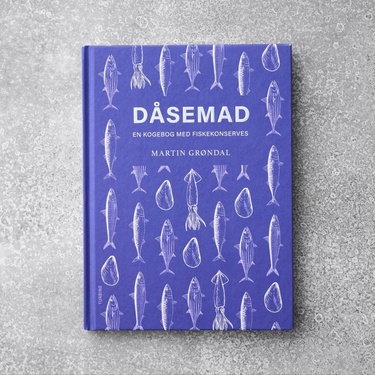 Den lilla kogebog "Kogebogen DÅSEMAD" fra Grøndals viser illustrationer af hvid fisk og skaldyr på omslaget, hvilende på en tekstureret grå overflade.