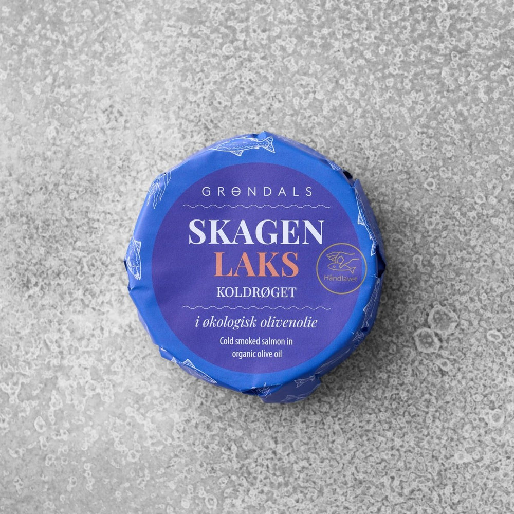 En rund pakke med Grøndals Koldrøget laks i økologisk olivenolie, pakket ind i blåt papir med hvid og guld tekst, placeret på en grå tekstureret overflade.