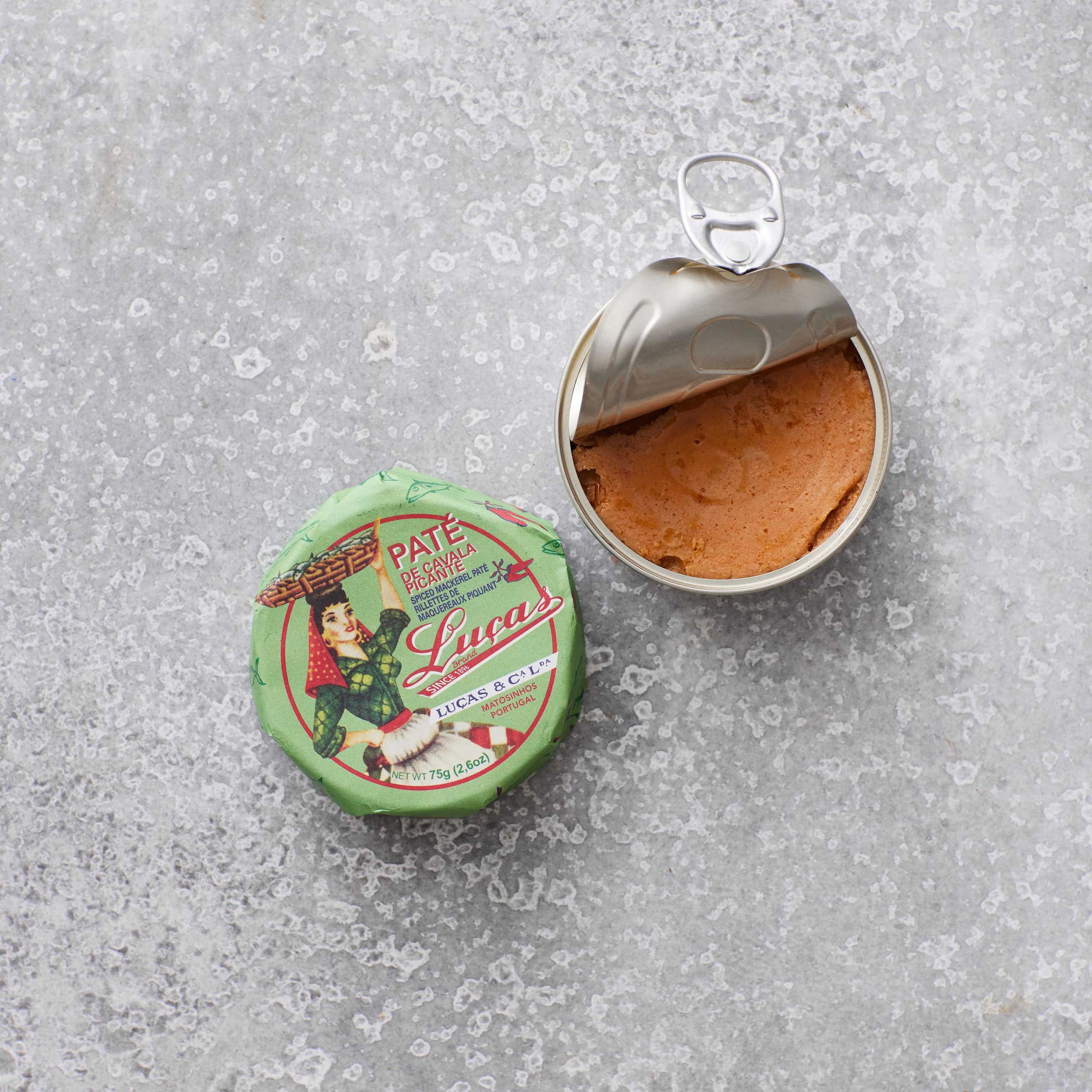 En åbnet dåse Lucas Spicy makrelpaté ligger på en grå, tekstureret overflade ved siden af det grønne låg, som har en illustration af en mand i traditionelt tøj, der holder et fad.