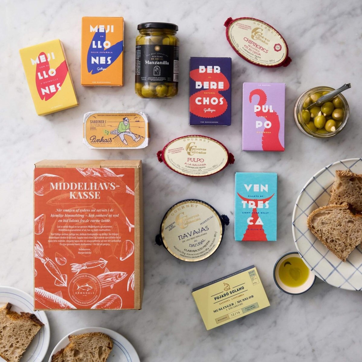 Grøndals’ Middelhavskassen byder på middelhavskonserverede fisk og skaldyr, herunder sardiner, samt oliven og brød – smukt arrangeret med farverig emballage og en glasskål med grønne oliven til den perfekte tapas-anretning.