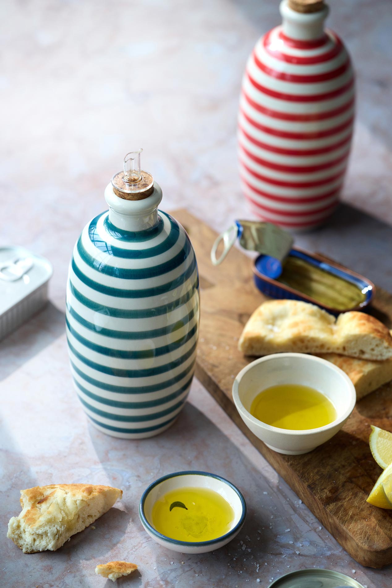 En grøn- og hvidstribet keramikflaske står ved siden af en lille skål med Grøndals Øko ekstrajomfru-olivenolie Arbequina-Picual (EVOO), brød og et træbræt. En rød- og hvidstribet flaske og mere brød ses i baggrunden.