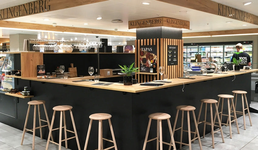 En moderne, minimalistisk tapasbar med en sort disk, træstole, glas og en lille plante. I baggrunden ses hylder med flasker og dagligvarer, og en person arbejder bag disken.