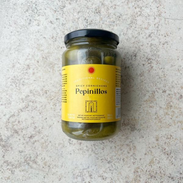 En glaskrukke med Spanske delikatesse cornichoner fra Don Gastronom med en gul etiket og sort låg er præsenteret på en lys, tekstureret overflade; perfekt til servering til tapas.