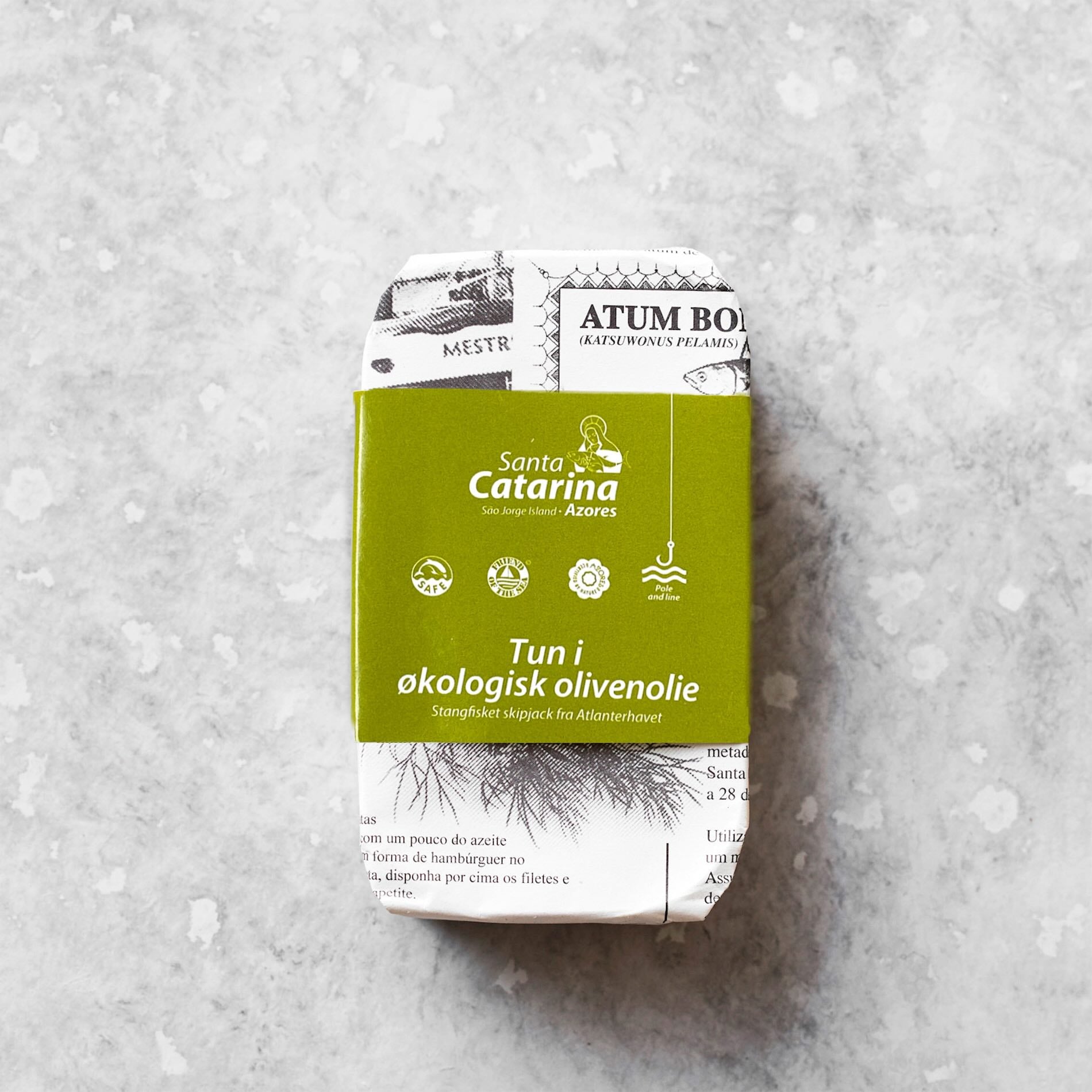 En dåse Santa Catarina Småstykker af tun i økologisk olivenolie, i grøn og avislignende emballage, står på en lysegrå plettet overflade.