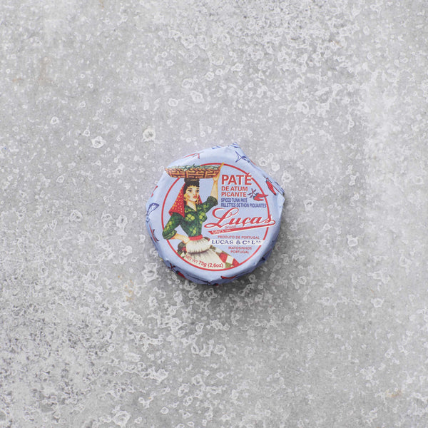 En lille rund dåse Lucas Spicy tunpaté med en vintage-etiket, der viser en kvinde, der bærer en kurv på hovedet, placeret på en tekstureret grå overflade.