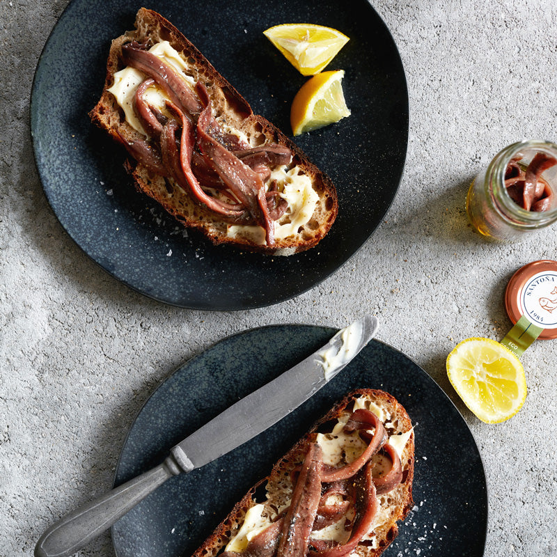 To skiver brød med smør og Pujado Solano OUTLET | Saltede ansjosfileter ØKO olivenolie på glas hviler på mørke tallerkener med citronbåde. Et åbent glas med ansjoserne og en smørkniv står på den grå strukturerede overflade.