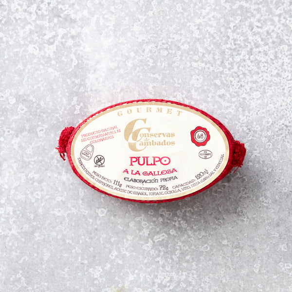 Oval dåse med Pulpo i galisisk sauce fra Conservas De Cambados med en fløde- og rød etiket med gourmetbranding. Dåsen indeholder mørt blækspruttearme i fyldig galisisk sauce, præsenteret på en tekstureret grå overflade med røde accenter.