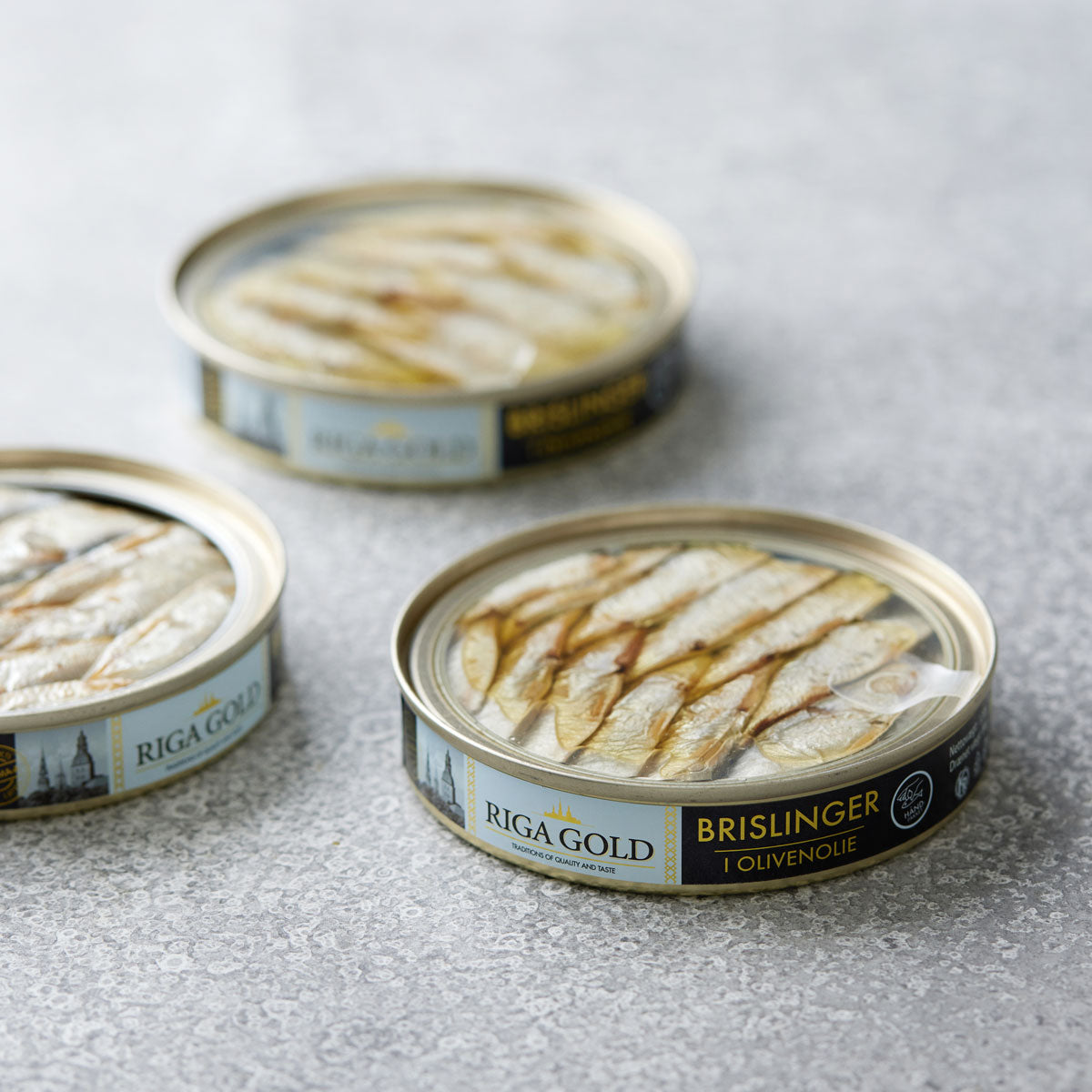 Tre åbne dåser Riga Gold Små brislinger i olivenolie er arrangeret på en tekstureret lysegrå overflade, med fisken pænt pakket og etiketterne synlige – en sand delikatesse blandt nordiske sardiner.