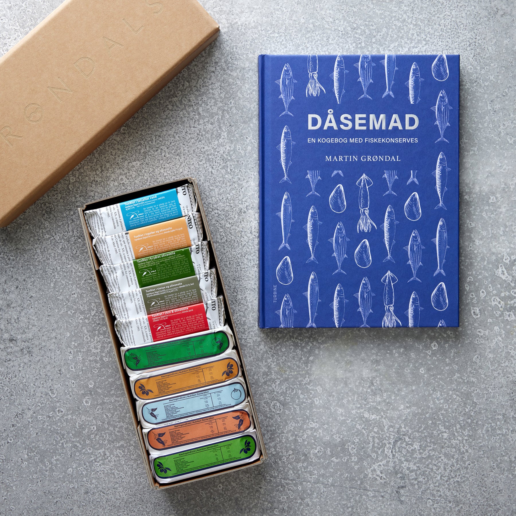 En blå “DÅSEMAD” kogebog med fiskeillustrationer ligger på en grå overflade ved siden af en farverig Gavekasse og DÅSEMAD-æske med Grøndals fiskekonserves. Et brunt paplåg er delvist synligt til venstre.
