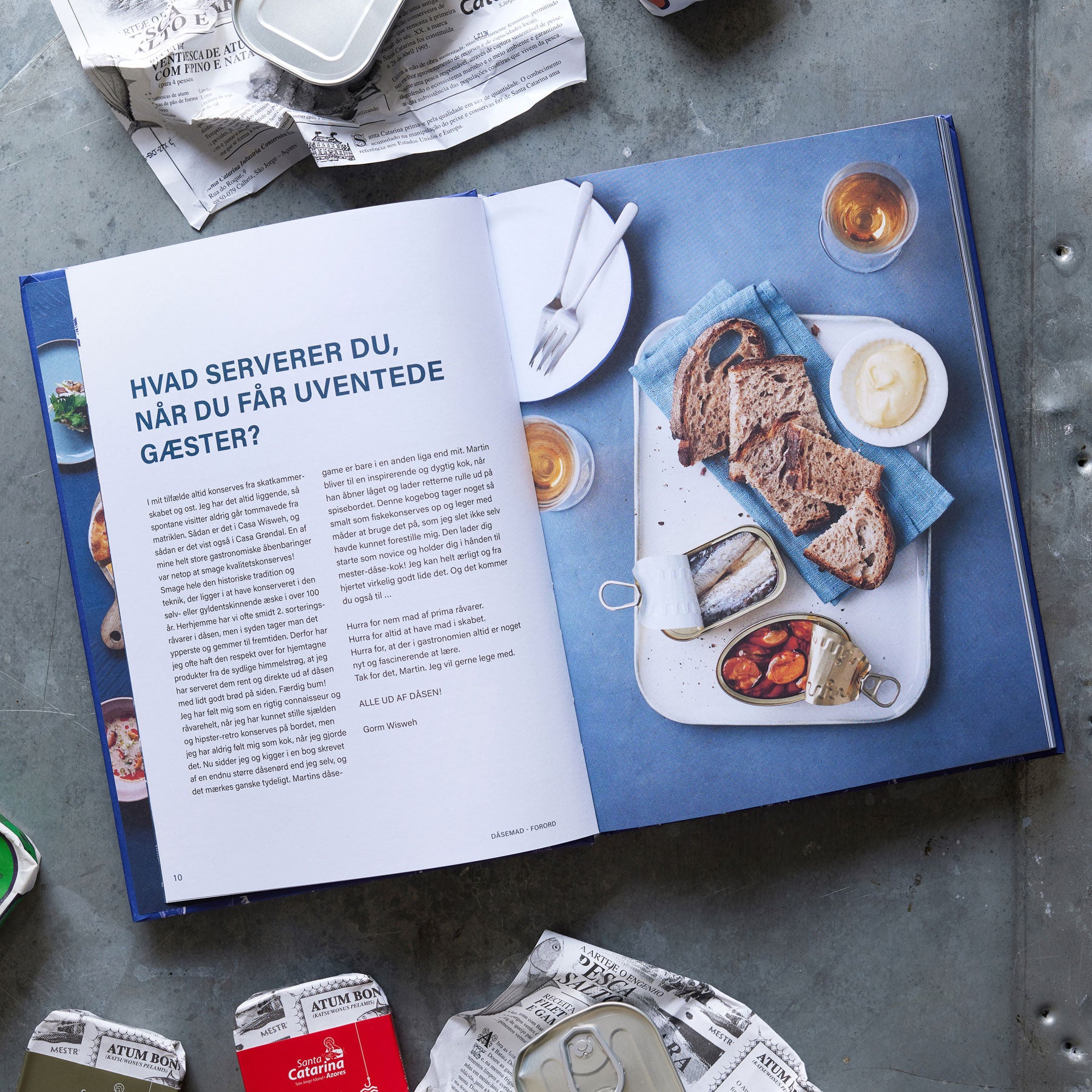 En åben kogebog på et gråt bord viser en dansk opskrift ved siden af Grøndals Smagekassen og DÅSEMAD: brød, citronbåde, dåsefisk, åbnede fiskekonservesdåser, papirpapir og krøllet avispapir fuldender scenen.