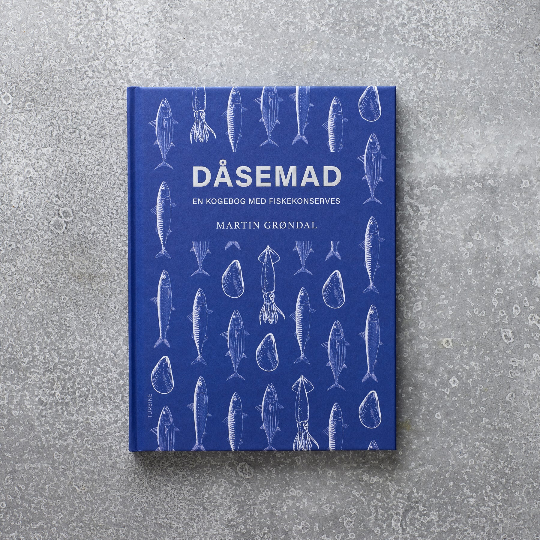 Bogen “Dåsemad - En kogebog med fiskekonserves” af Martin Grøndal, med en blå forside dekoreret med hvide illustrationer af forskellige fisk og skaldyr, set mod en grå tekstureret baggrund.⬤