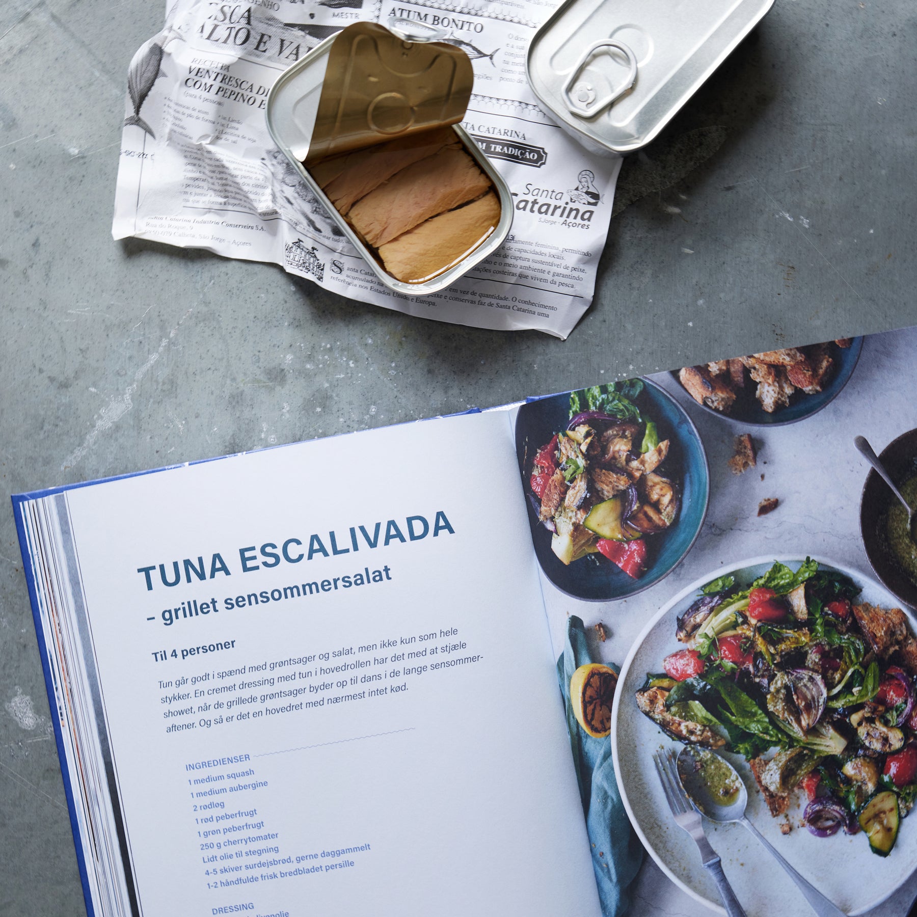 En åben kogebog med en opskrift på Tuna Escalivada ligger ved siden af en salat og en åbnet dåse fra Gavekasse og DÅSEMAD fiskekonserves på avispapir, alt sammen anrettet på en grå bordplade.