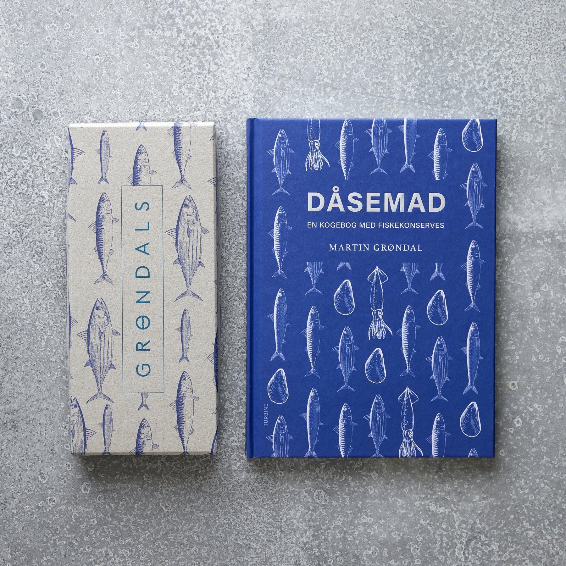 En blå DÅSEMAD kogebog med fiskeillustrationer og en beige Grøndals Værtsgave-æske, begge dekoreret med fiskekonserves-tegninger, er placeret på en spættet grå overflade.