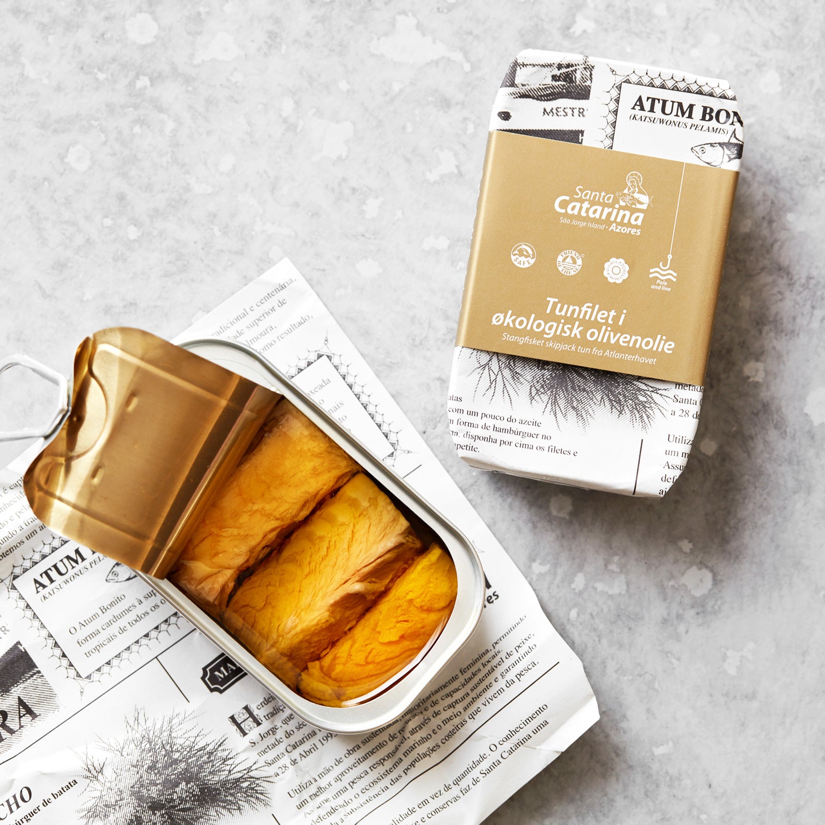 En åbnet dåse Santa Catarina Tunfilet i økologisk olivenolie ligger på en avis, mens en anden beige-indpakket dåse af det samme produkt ligger i nærheden på en lys, tekstureret overflade.