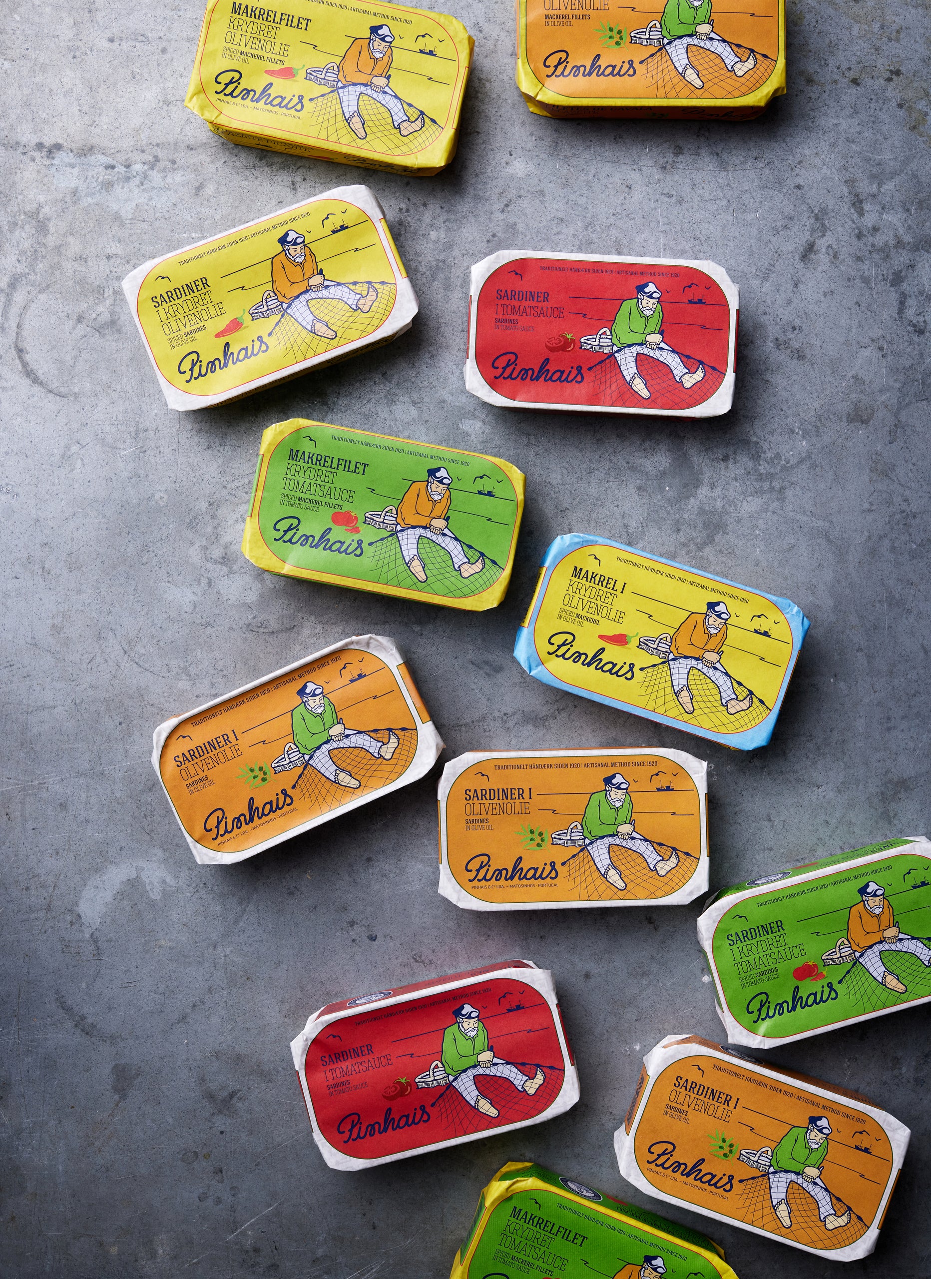 Forskellige farverige dåser med Pinhais-sardiner med illustrerede fiskemotiver er arrangeret på en grå, struktureret overflade. Dåserne er røde, gule, orange, grønne og blå.