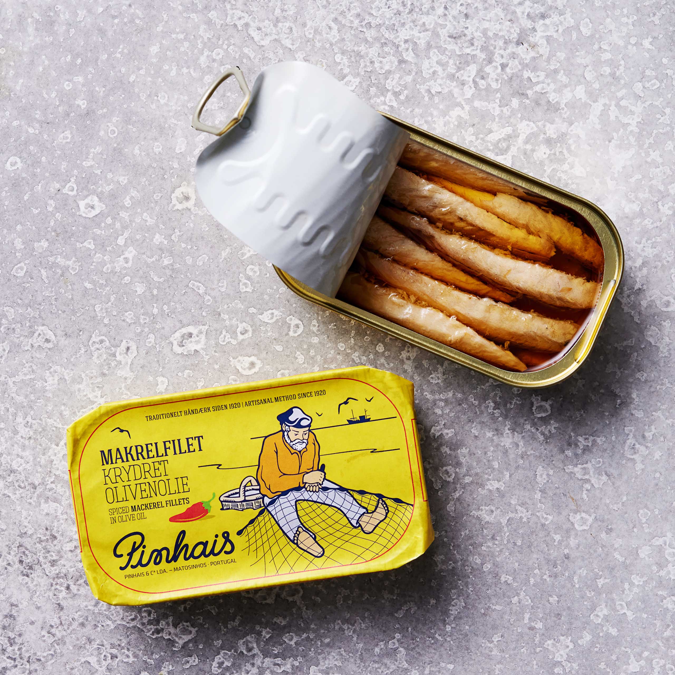 En åbnet dåse Pinhais Makrelfilet i krydret olivenolie hviler på en grå overflade ved siden af det gule låg med en illustration af en fisker og dansk og engelsk tekst, der fremhæver bæredygtigt fiskeri.